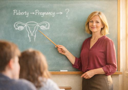 Confident midlife woman teaching about ovaries and menopause in a classroom, pointing to a chalkboard showing “Puberty → Pregnancy → ?”