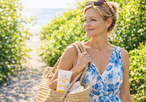 Woman over 40 wearing sunglasses and a summer dress carrying a beach bag with SPF 50 sunscreen visible