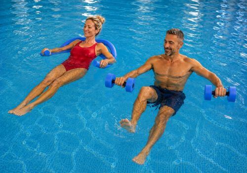 Middle-aged woman floating with a pool noodle while a man performs Aqua Pilates with foam dumbbells, demonstrating how buoyancy supports the body in water.
