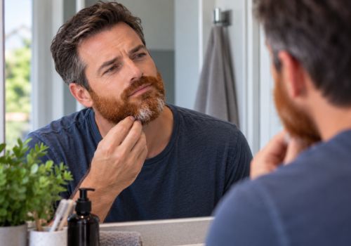 Man with dark hair examining his reddish beard with gray hairs in the mirror
