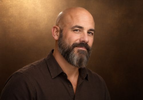 Bald man with a shaped medium-length salt-and-pepper beard and tapered sideburns against a bronze-brown background.